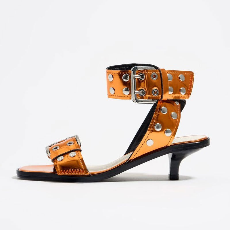Metallic orange patent leather strappy mid-heel sandal with silver rivets, an adjustable buckle, and a black kitten heel.