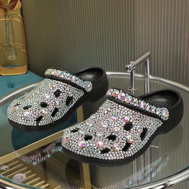 Black slide sandals encrusted with iridescent rhinestones and large crystals on a glass table.
