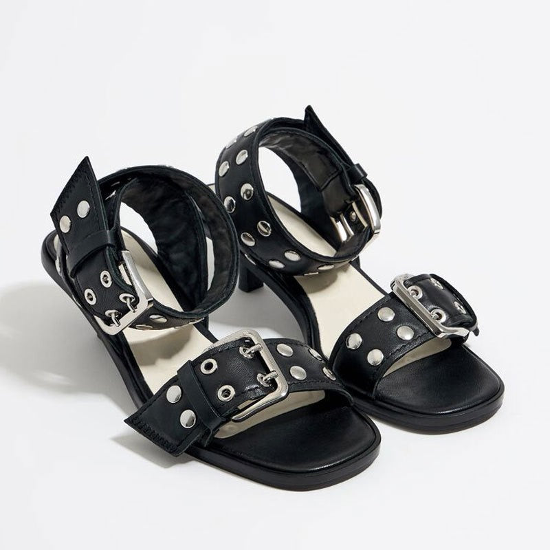 Black faux-leather strappy mid-heel sandals with silver rivet studs and large metallic buckles on ankle and toe straps.