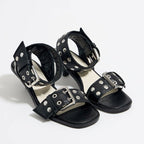 Black faux-leather strappy mid-heel sandals with silver rivet studs and large metallic buckles on ankle and toe straps.