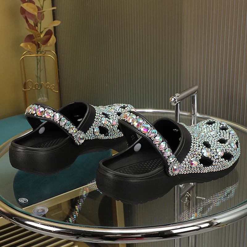 Black platform clogs featuring iridescent rhinestone embellishments and a pivoting heel strap on a glass table.