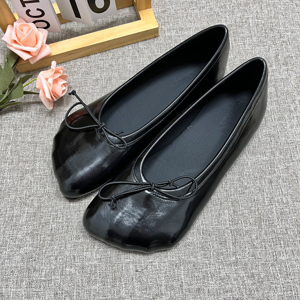 Sophisticated black glossy slip-on flats with round toes and decorative front bows on a grey textured background.