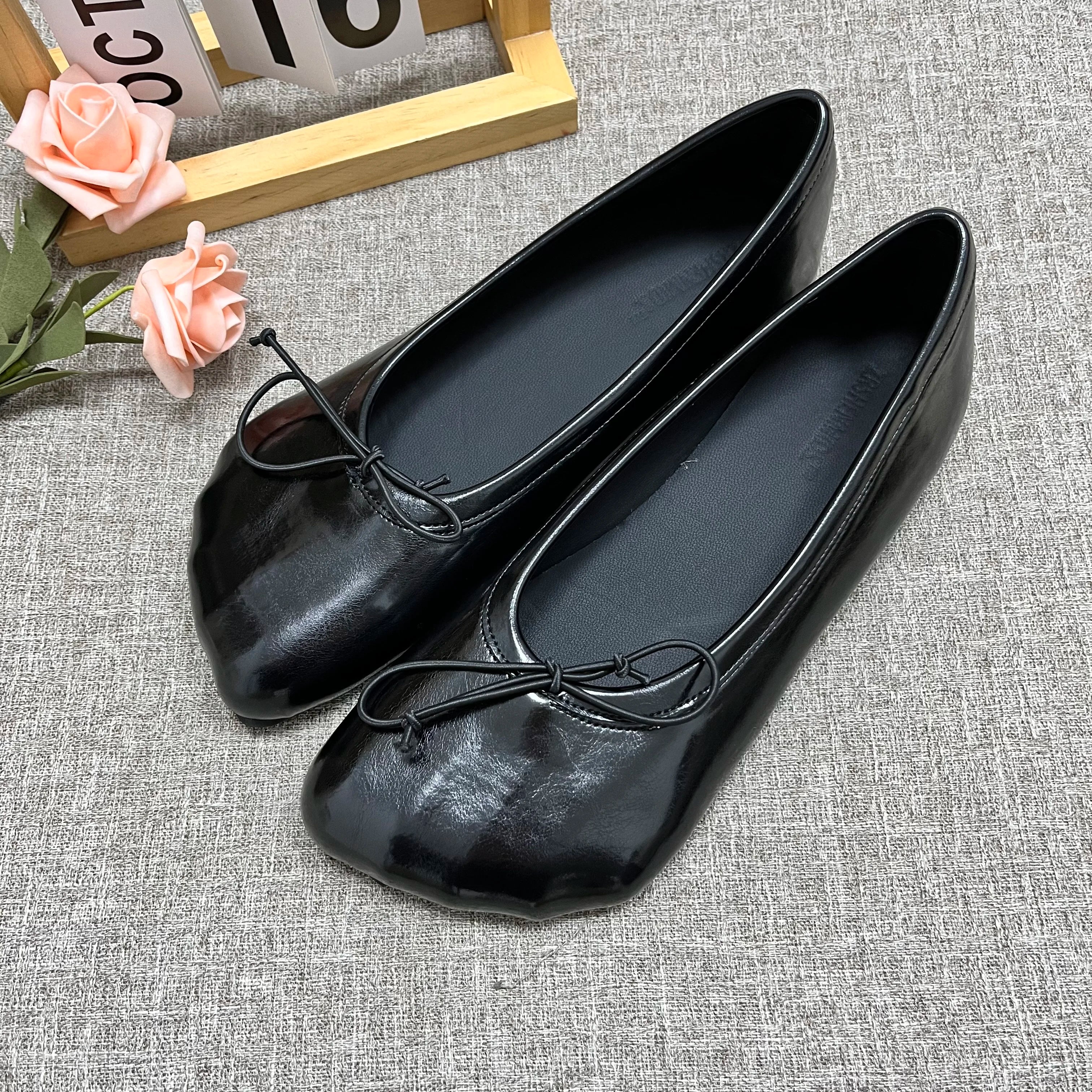 Sophisticated black glossy slip-on flats with round toes and decorative front bows on a grey textured background.
